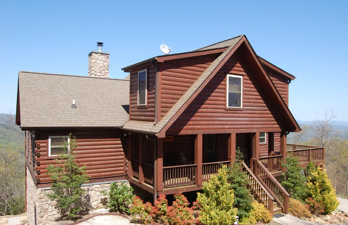 A welcoming exterior view of the Cedar Rock Cabin, showcasing its classic log cabin structure with a stone chimney, lush green landscaping, and an inviting porch with railings. The scene evokes a sense of arrival and highlights outdoor spaces perfect for gatherings and relaxation.