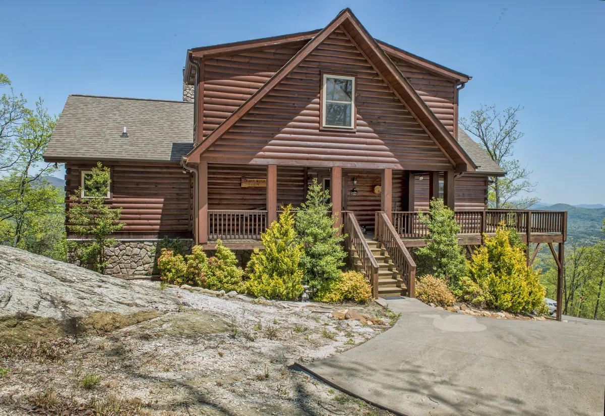 The rustic log cabin sits atop rocky terrain, surrounded by a landscaped garden. The front porch is visible, suggesting the perfect spot to relax while taking in the mountain scenery. The overall mood is cozy and serene, with hints of outdoor activity and peaceful family gatherings.