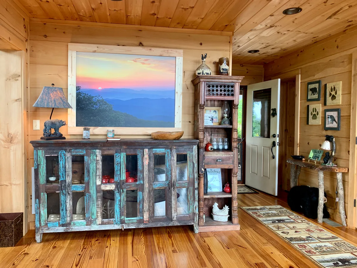 A cozy living area with a rustic wooden cabinet, mountain view artwork, decorative shelves, and an open door leading outside. The space feels welcoming, ideal for quiet moments, reading, or gathering with friends, and hints at direct access to fresh air and nature.