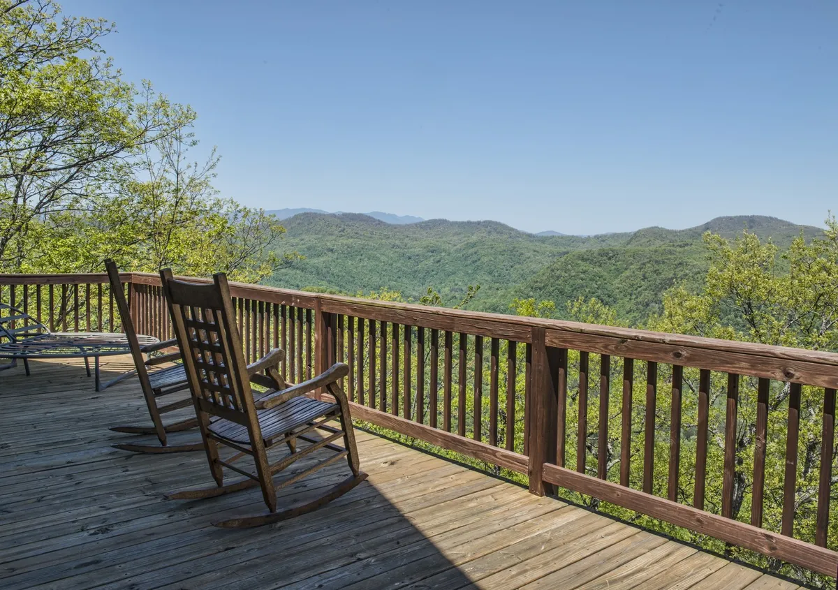 A serene corner of the wraparound deck featuring classic wooden rocking chairs overlooking a sweeping mountain vista. The deck's wooden railings provide a sense of safety without blocking the panoramic views, and the tranquil setting encourages relaxation and quiet moments.