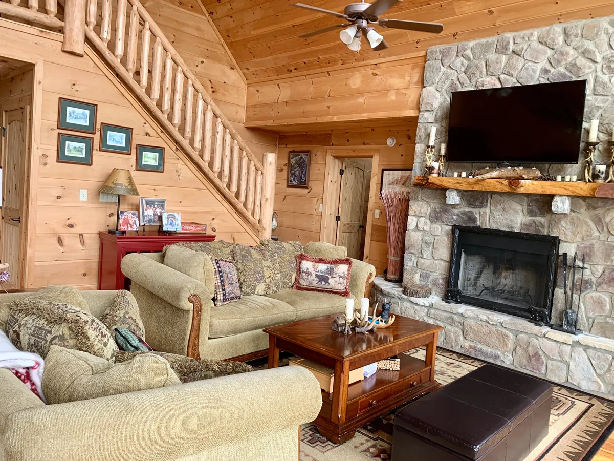 A welcoming great room featuring a prominent stone fireplace surrounded by plush couches and a sturdy wooden coffee table. Wooden beams overhead and ample natural light create a warm, inviting atmosphere—the perfect spot for family gatherings or relaxing by the fire.