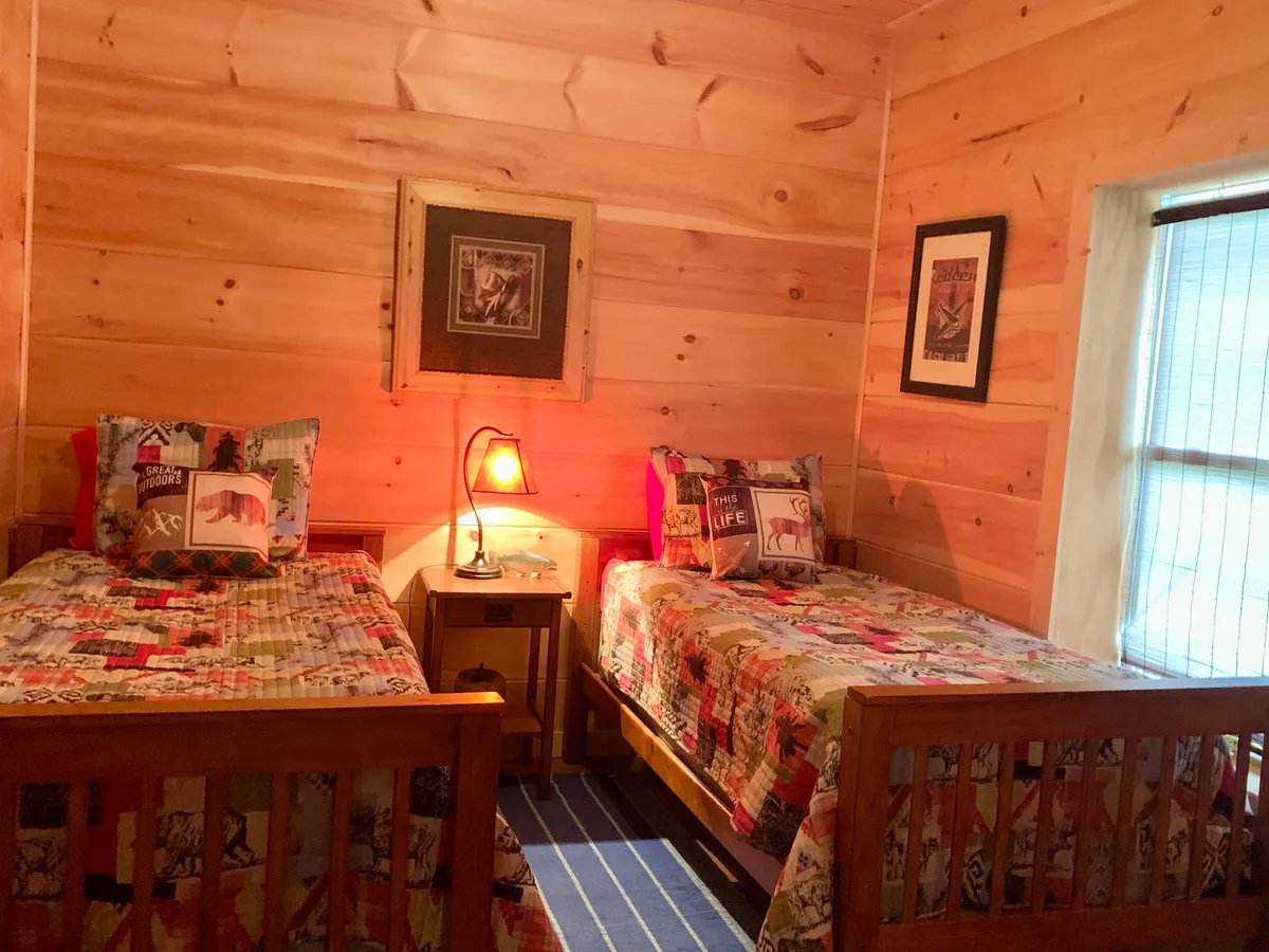 A charming twin bedroom with two wooden beds, each topped with colorful, patterned quilts. A shared nightstand with a lamp sits between the beds, and framed artwork adorns the wall, creating a playful yet homey setting ideal for children or friends.