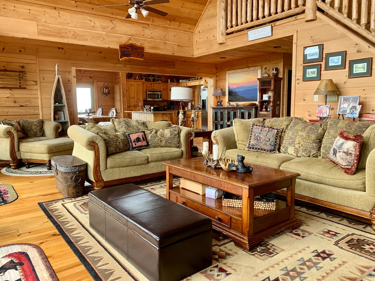 A spacious, warmly lit living room with comfortable sofas, a patterned rug, wooden coffee table, and a wooden ceiling. The area is perfect for group gatherings, reading, or socializing, and the large windows suggest beautiful views and plenty of natural light.