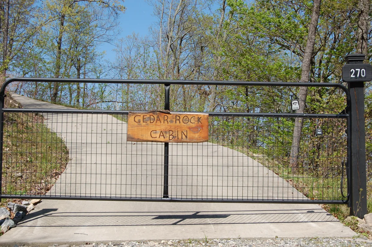 A welcoming entrance to Cedar Rock Cabin, featuring the distinctive cabin sign, a rustic gate, and a winding driveway that leads up through wooded terrain. The image suggests a sense of discovery, emphasizing both the journey and the privacy of the setting.