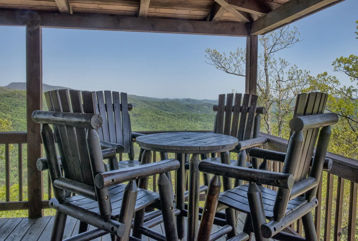 Wooden chairs and a round table on the deck with a breathtaking mountain backdrop. The casual arrangement suggests gatherings for coffee, meals, or evening drinks while enjoying the scenery. The open-air setting captures the essence of al fresco dining.