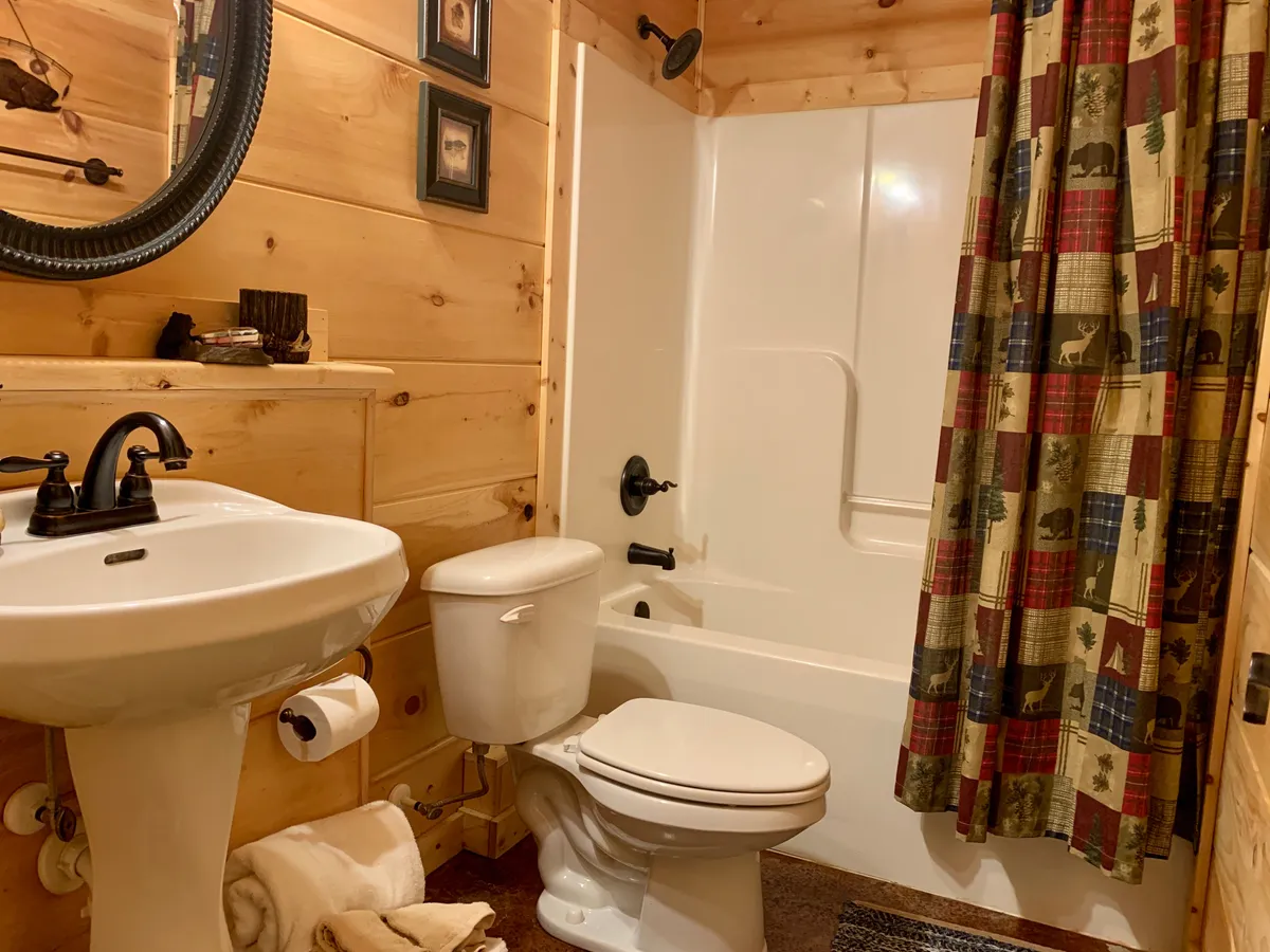 A bathroom with a pedestal sink, a cute plaid shower curtain over a shower area, a decorative mirror, and rustic wooden walls. The space feels clean, functional, and inviting, echoing the cabin’s blend of comfort and style.