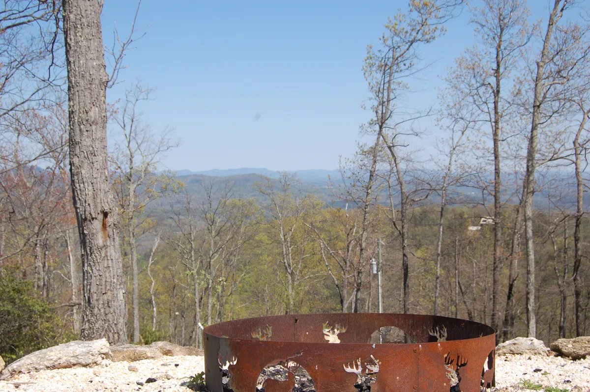 An inviting outdoor fire pit surrounded by natural mountain scenery, bordered by trees and overlooking distant peaks. The setting is perfect for evening gatherings, s’mores, and storytelling under the stars.