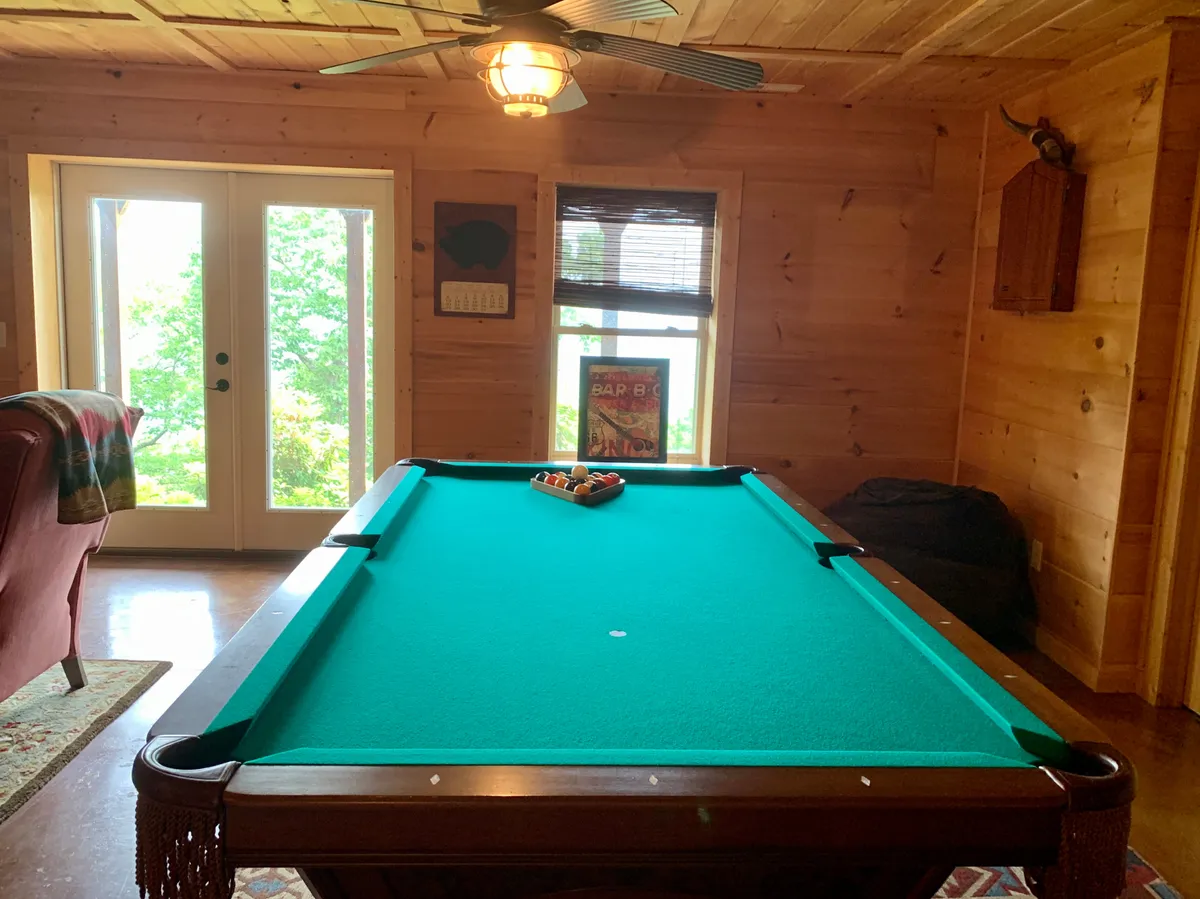 A vibrant downstairs family room featuring a pool table at the center, large windows that offer natural light, and comfortable seating with decorative wall elements. The overall layout suggests a lively yet welcoming space perfect for games, socializing, and relaxing together.