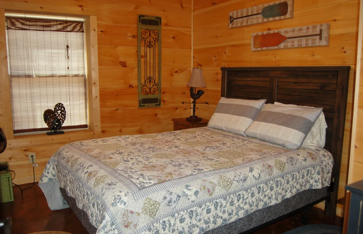 A warmly lit bedroom featuring a bed with a cozy quilt, a sturdy wooden headboard, and a lamp on the nightstand. Sunlight filters softly through a nearby window, illuminating the inviting textures of the bedding and creating a restful atmosphere.
