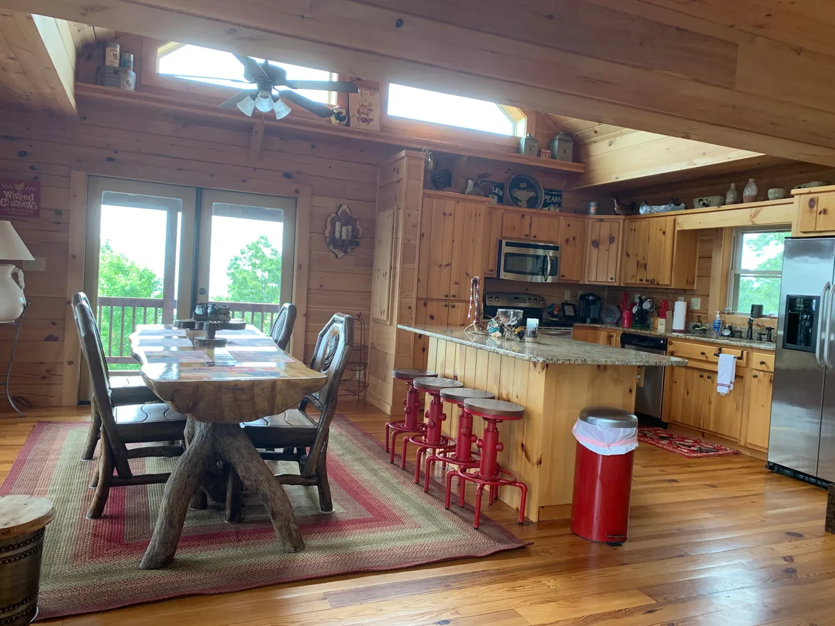 A spacious dining area with a rustic wooden dining table and chairs, a kitchen island, and large windows allowing sunlight to flood the space. The open floor plan unites dining and kitchen areas, perfect for meals, socializing, and enjoying expansive views.