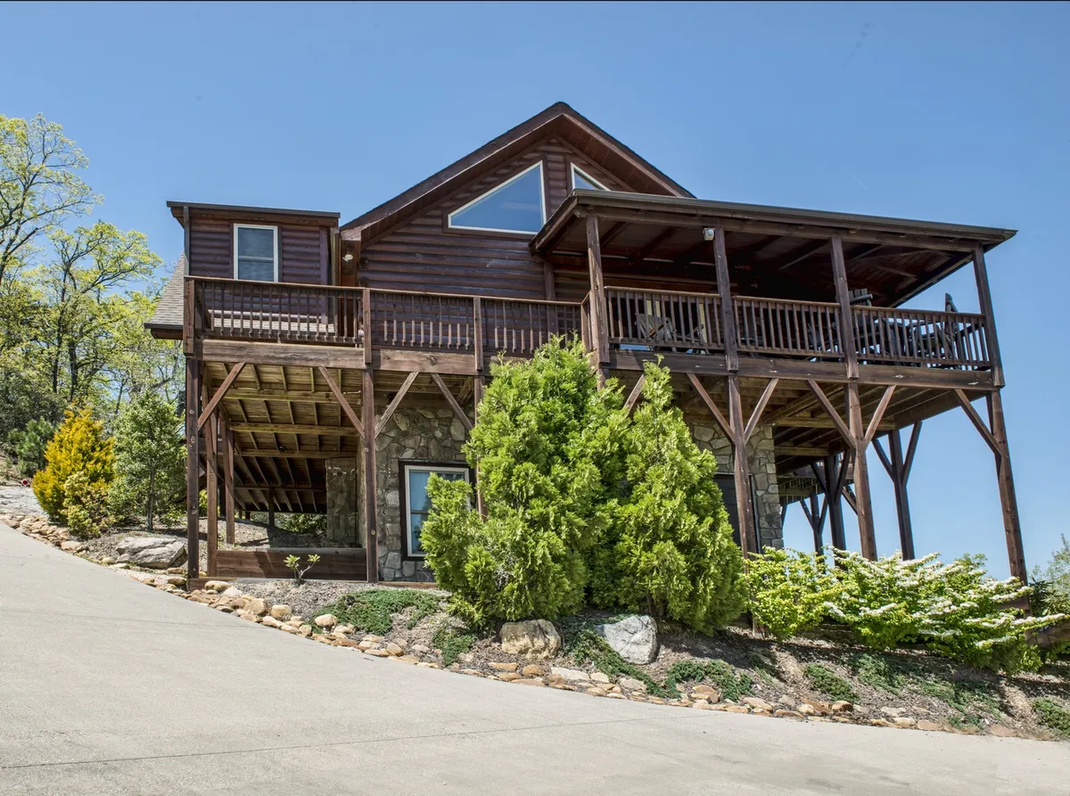 A vibrant exterior shot featuring a wooden balcony overlooking lush greenery, a stone facade, and an angled roof. The setting is ideal for outdoor dining, stargazing, and family barbecues, with space for group activities and relaxation.