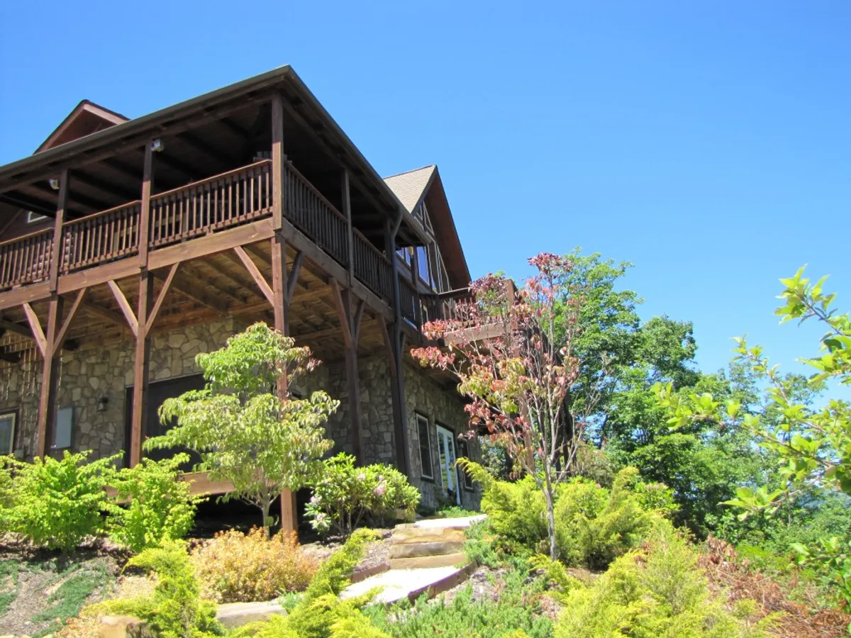 A welcoming mountain cabin with a spacious wooden balcony overlooking vibrant landscaping and panoramic views, featuring a stunning stone exterior; ideal for relaxing, outdoor gatherings, and sunset watching.